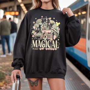 Może przedstawiać: Czarna bluza z kolorowym nadrukiem graficznym przedstawiającym zamek, filiżankę i inne ilustracje. Tekst "The Most Magical Place" jest wyśrodkowany pod grafiką. Bluza ma długie rękawy i okrągły dekolt.
