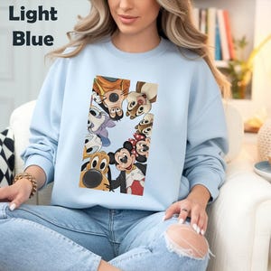 May include: Light blue sweatshirt featuring a rectangular graphic of various cartoon characters, including Mickey and Minnie Mouse, Goofy, and Pluto. The text "Light Blue" is in the upper left corner. The sweatshirt is worn by a person.