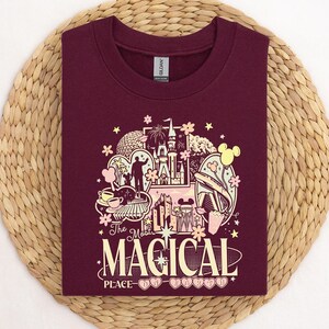Może przedstawiać: Bordowa bluza z kolorową grafiką przedstawiającą zamek, Myszka Miki i napis "The Most Magical Place on Earth". Bluza wykonana jest z miękkiego materiału.