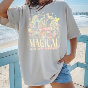 Może przedstawiać: Kremowa koszulka z kolorową grafiką zamku, uszami Myszki Miki i innymi obrazami z parku rozrywki. Tekst "The Most MAGICAL PLACE on Earth" jest wydrukowany poniżej. Koszulka jest noszona na plaży.