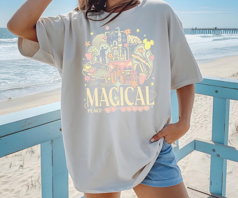 Może przedstawiać: Kremowa koszulka z kolorową grafiką zamku, uszami Myszki Miki i innymi obrazami z parku rozrywki. Tekst "The Most MAGICAL PLACE on Earth" jest wydrukowany poniżej. Koszulka jest noszona na plaży.