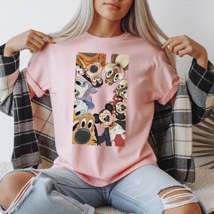 May include: A light pink t-shirt featuring a rectangular graphic of Disney cartoon characters, including Mickey Mouse, Minnie Mouse, Donald Duck, and Goofy. The characters are arranged in a circle, with their faces visible. The shirt has short sleeves.