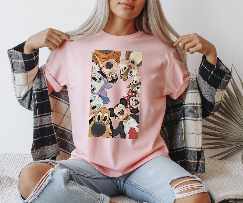 May include: A light pink t-shirt featuring a rectangular graphic of Disney cartoon characters, including Mickey Mouse, Minnie Mouse, Donald Duck, and Goofy. The characters are arranged in a circle, with their faces visible. The shirt has short sleeves.