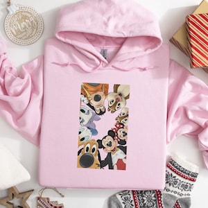 May include: A light pink hooded sweatshirt featuring a colorful graphic of various cartoon characters. The graphic includes Mickey Mouse, Pluto, and other Disney characters. The sweatshirt is made of a soft material and has a drawstring hood.