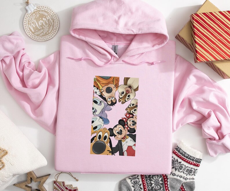 May include: A light pink hooded sweatshirt featuring a colorful graphic of various cartoon characters. The graphic includes Mickey Mouse, Pluto, and other Disney characters. The sweatshirt is made of a soft material and has a drawstring hood.