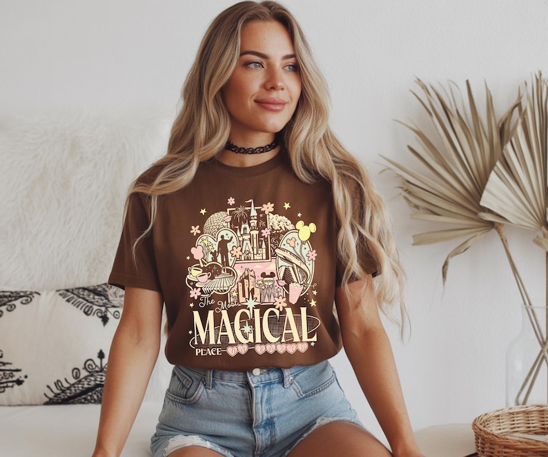 Może przedstawiać: Brązowa koszulka z kolorowym nadrukiem graficznym. Projekt zawiera ilustracje zamku, kwiat&oacute;w i innych fantazyjnych element&oacute;w z napisem "The Magical Place". Koszulka wykonana jest z miękkiego materiału.