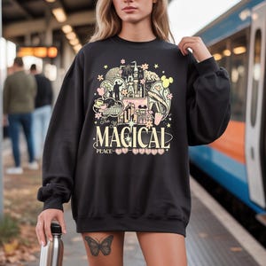 May include: Black sweatshirt with a colourful graphic design featuring a castle, teacup, and other illustrations. The text "The Most Magical Place" is centered below the graphic. The sweatshirt has long sleeves and a crew neck.