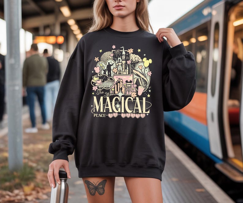 May include: Black sweatshirt with a colourful graphic design featuring a castle, teacup, and other illustrations. The text "The Most Magical Place" is centered below the graphic. The sweatshirt has long sleeves and a crew neck.