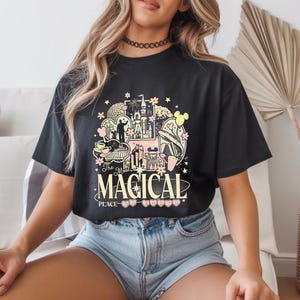 Może przedstawiać: Czarny t-shirt z kolorowym wzorem graficznym przedstawiającym zamek, uszy Myszki Miki i napis "The Most Magical Place". Projekt zawiera r&oacute;żowe, ż&oacute;łte i zielone elementy. Koszulka jest noszona przez osobę.