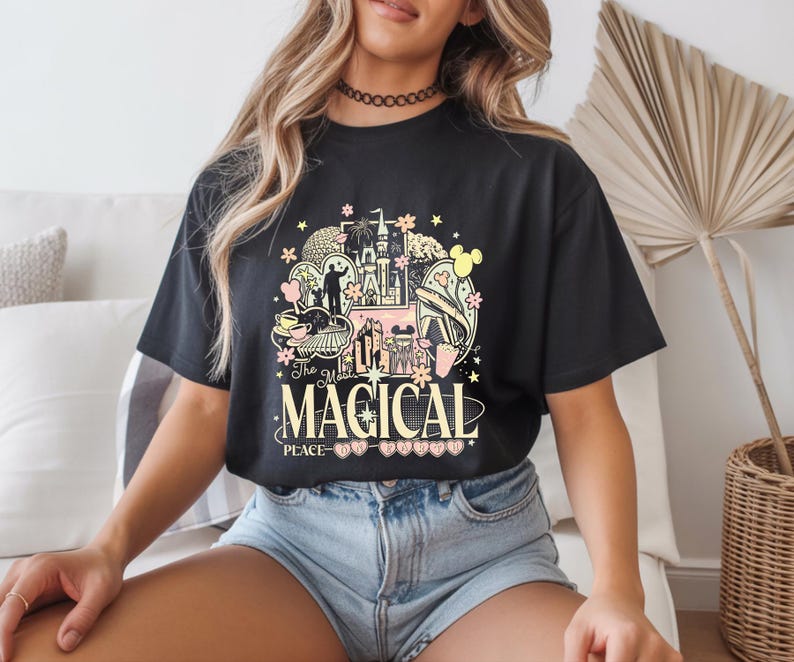 Może przedstawiać: Czarny t-shirt z kolorowym wzorem graficznym przedstawiającym zamek, uszy Myszki Miki i napis "The Most Magical Place". Projekt zawiera r&oacute;żowe, ż&oacute;łte i zielone elementy. Koszulka jest noszona przez osobę.