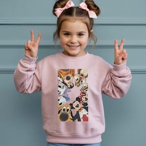 May include: A light pink sweatshirt featuring a colorful graphic of Disney cartoon characters. The graphic includes Mickey Mouse, Minnie Mouse, Goofy, and Chip and Dale. The sweatshirt is worn by a young child.