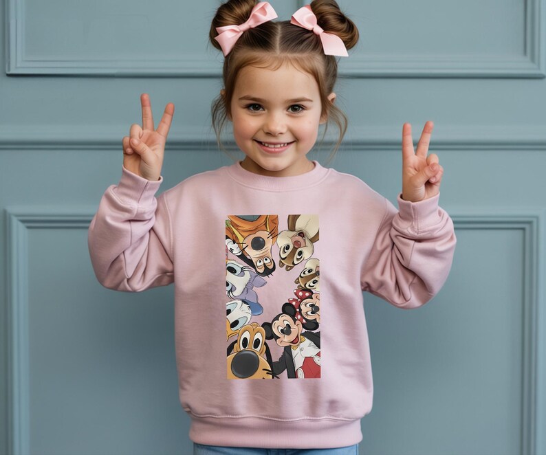 May include: A light pink sweatshirt featuring a colorful graphic of Disney cartoon characters. The graphic includes Mickey Mouse, Minnie Mouse, Goofy, and Chip and Dale. The sweatshirt is worn by a young child.
