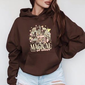 Może przedstawiać: Brązowa bluza z kapturem z kolorową grafiką z napisem "The Most Magical Place". Grafika zawiera zamek, uszy Myszki Miki i inne fantazyjne elementy. Bluza ma przednią kieszeń.