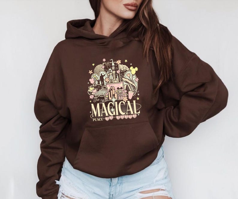 Może przedstawiać: Brązowa bluza z kapturem z kolorową grafiką z napisem "The Most Magical Place". Grafika zawiera zamek, uszy Myszki Miki i inne fantazyjne elementy. Bluza ma przednią kieszeń.