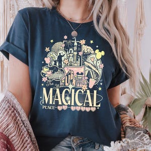 Może przedstawiać: Granatowa koszulka z kolorową grafiką przedstawiającą zamek, Myszka Miki i napis "The Most Magical Place". Projekt zawiera pastelowe r&oacute;żowe, ż&oacute;łte i niebieskie elementy. Koszulka ma podwinięte rękawy.