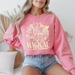 Może przedstawiać: R&oacute;żowa bluza z nadrukiem przedstawiającym zamek, uszy Myszki Miki i napis "The Most Magical Place". Projekt jest w odcieniach ż&oacute;łci i r&oacute;żu. Bluza ma długie rękawy.