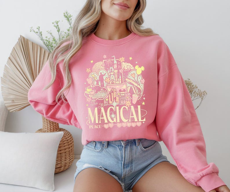 Może przedstawiać: R&oacute;żowa bluza z nadrukiem przedstawiającym zamek, uszy Myszki Miki i napis "The Most Magical Place". Projekt jest w odcieniach ż&oacute;łci i r&oacute;żu. Bluza ma długie rękawy.