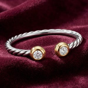 May include: A silver and gold bracelet with a twisted design. The bracelet features two gold-colored, circular settings, each holding a sparkling, clear gemstone. The jewelry rests on a deep red velvet surface.