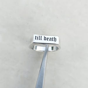 Könnte beinhalten: Ein silberner Ring mit dem Schriftzug "till death" in schwarzer gotischer Schrift. Der Ring wird von einer silbernen Pinzette vor weißem Hintergrund gehalten. Der Ring hat eine rechteckige Oberfläche.