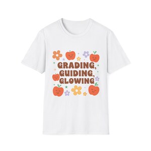 May include: White t-shirt with a colorful graphic design. The design features orange apples, flowers, and the words "GRADING, GUIDING, GLOWING" in brown letters. The shirt is a casual, everyday wear item.