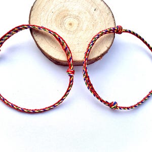 Puede incluir: Dos pulseras de amistad hechas a mano. Cada pulsera está hecha de hilos trenzados rojos, amarillos, verdes y azules. Las pulseras están anudadas en los extremos y se exhiben sobre una rodaja de madera.