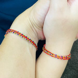 Könnte beinhalten: Zwei geflochtene Freundschaftsarmbänder, eines an jedem Handgelenk, sind im Bild zu sehen. Die Armbänder bestehen aus roten, gelben, blauen und weißen Fäden. Die Handgelenke sind vor einem dunkelblauen und grauen Hintergrund.