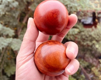 May include: Two smooth, spherical wooden balls with a rich reddish-brown color are held in a hand. The balls have a polished finish, showcasing the wood grain. The background is blurred, with green foliage.