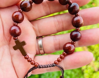 May include: A dark brown wooden rosary bracelet with large, round beads and a small cross charm. The bracelet is held in a hand, showcasing the natural wood grain and the intricate knotting of the adjustable cord.