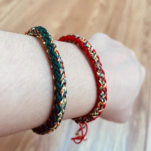 Può includere: Vengono mostrati due braccialetti intrecciati, uno verde e uno rosso. Il braccialetto verde ha accenti gialli, arancioni e bianchi. Il braccialetto rosso ha accenti simili. Entrambi sono indossati su un polso.