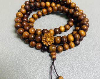Op de afbeelding: Een armband met meerdere strengen, bestaande uit talloze kleine, ronde, donkerbruine houten kralen. De armband is voorzien van een centrale, uit hout gesneden bloemhanger en een koordverlenging afgewerkt met extra bijpassende kralen.