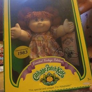 Peut inclure: Poupée Cabbage Patch Kids vintage dans sa boîte d'origine. La poupée a des cheveux roux en fil et porte une robe à motifs. La boîte est jaune et verte, avec le texte "Commémoration 1983" et "Édition Vintage Limitée". La boîte indique également "Je suis spécial et unique. Je suis adoptable !"