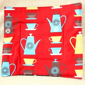 May include: A square quilted fabric potholder in bright red featuring a repeating pattern of stylized teapots and teacups in shades of light blue, yellow, and brown. The teapots are decorated with a central starburst design on their sides.