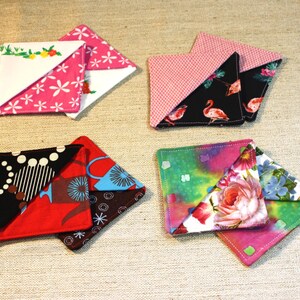 May include: Four sets of square fabric corner bookmarks featuring diverse patterns. One set has pink and white floral prints, another features black fabric with pink flamingos, a third displays red and black geometric designs, and the last shows floral motifs.