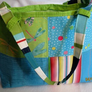 May include: A patchwork tote bag with a variety of colorful patterns. The bag features a green dragonfly print, polka dots, and stripes. The shoulder strap is a mix of colors. The bag is approximately 14 inches wide and 12 inches tall.