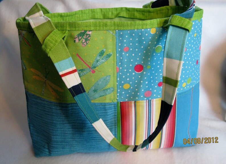 May include: A patchwork tote bag with a variety of colorful patterns. The bag features a green dragonfly print, polka dots, and stripes. The shoulder strap is a mix of colors. The bag is approximately 14 inches wide and 12 inches tall.