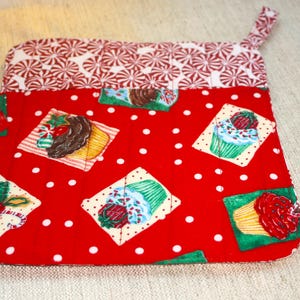May include: A red and white quilted oven mitt with a loop for hanging. The mitt features a red background with white polka dots and cupcake designs. The top section has a pattern of red and white peppermint candies.