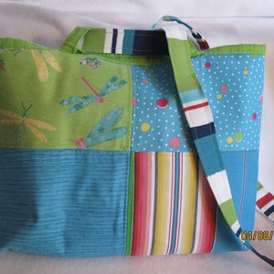 May include: A patchwork tote bag with a variety of colorful patterns. The bag features panels of green with dragonfly designs, blue with white polka dots, and striped fabric. The strap is a mix of blue, white, and green stripes.