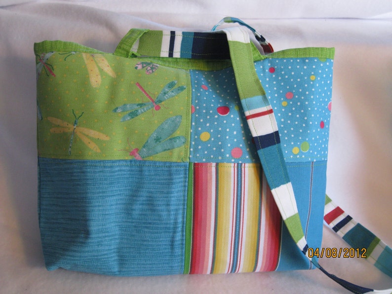 May include: A patchwork tote bag with a variety of colorful patterns. The bag features panels of green with dragonfly designs, blue with white polka dots, and striped fabric. The strap is a mix of blue, white, and green stripes.
