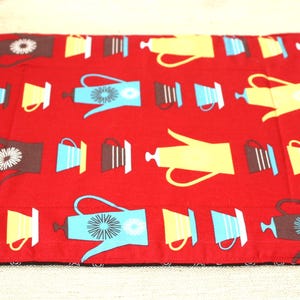 May include: A rectangular fabric placemat in a vibrant red color featuring a repeating pattern of stylized coffee pots and cups in shades of brown, light blue, and yellow. The design includes various coffee-related vessels arranged in rows.