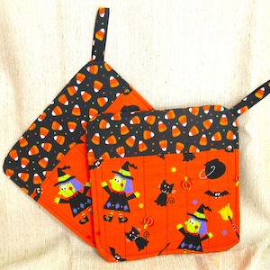May include: Two Halloween-themed pot holders. The top portion is black with candy corn. The bottom portion is orange with witch and black cat graphics. Each pot holder has a loop for hanging.