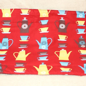 May include: A rectangular fabric placemat featuring a vibrant red background with a repeating pattern of stylized teapots and teacups in shades of yellow, light blue, and brown. The design includes various teapot shapes and matching cups on saucers.
