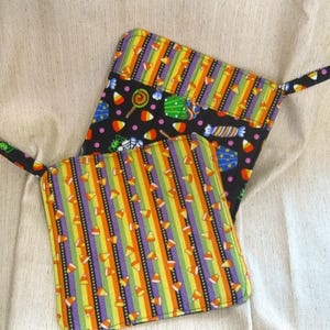 May include: Two Halloween-themed pot holders. One features a black background with candy and lollipop designs. The other has vertical stripes of orange, yellow, green, purple, and black with candy corn. Both have a small loop for hanging.