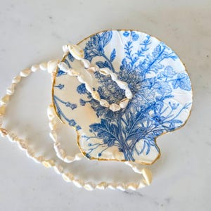 May include: A seashell-shaped dish with a blue floral design and gold trim, paired with a necklace made of small seashells. The dish features a detailed botanical print, creating a coastal and decorative theme.