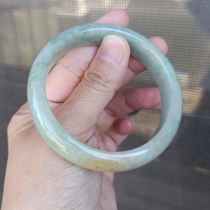 Peut inclure: Un bracelet jonc en jade circulaire et lisse, présentant une surface polie aux tons vert pâle translucides avec une zone distincte de couleur jaune doré chaud intégrée dans la pierre naturelle de cet anneau solide et continu.