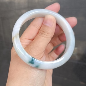 May include: A smooth, circular bangle bracelet crafted from translucent white stone with subtle, natural inclusions of deep green and pale brown. The polished surface reflects light, highlighting the rounded profile and the organic color variations.