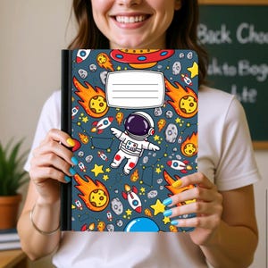 May include: A rectangular notebook with a dark blue cover featuring a repeating space pattern of an astronaut, rockets, yellow stars, orange comets, and gray asteroids. A white rectangular label with four horizontal lines is centered on the front cover.