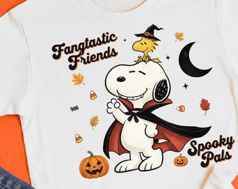 May include: White t-shirt featuring a Halloween-themed graphic. The design includes Snoopy in a vampire cape, Woodstock in a witch hat, pumpkins, candy corn, and the phrases "Fangtastic Friends" and "Spooky Pals."