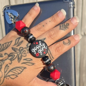 Puede incluir: Un dije de teléfono con cuentas negras y rojas con las palabras "good girl" y un gráfico de labios. El dije está sujeto a una funda de teléfono transparente con un cierre plateado en forma de corazón.