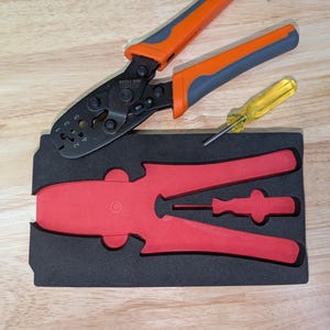 May include: A set of tools including a black and orange crimping tool, a yellow screwdriver, and a red tool set in a black foam insert. The tools are arranged on a light brown wooden surface.