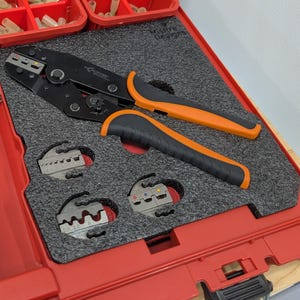 May include: Black and orange crimping tool with a black and grey foam insert, and a red storage case. The tool has orange handles and is designed for electrical work. Additional crimping dies are included.